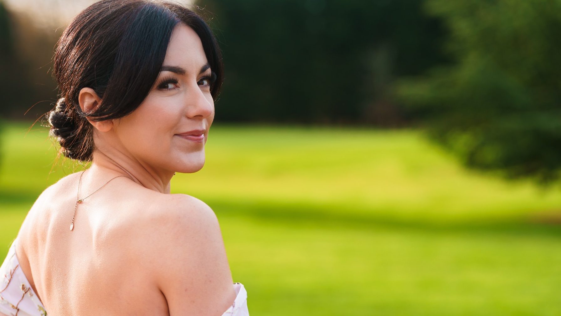 A bride on the lawn on her wedding day, looking back over her shoulder, makeup by Emily